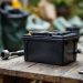 Batterie de tondeuse posée sur un établi de jardin, avec des outils d’entretien à proximité et des sacs de recyclage en arrière-plan.